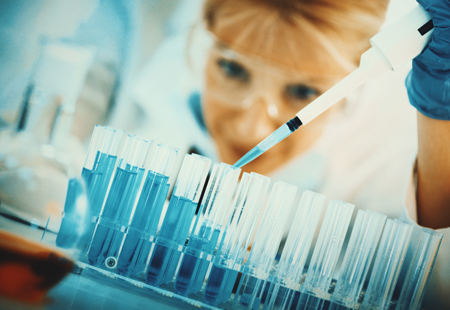 Closeup low angle view of mid 30's blond female chemist carefully measuring chemicals in a laboratory. Wearing protective mask and eyeglasses. She's using pipette to accurately put blue chemical into flasks.